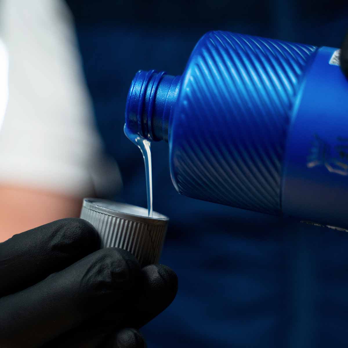 A person in a black glove pouring the clear, viscous Labocosmetica Revitax shampoo from its blue bottle into the silver cap.