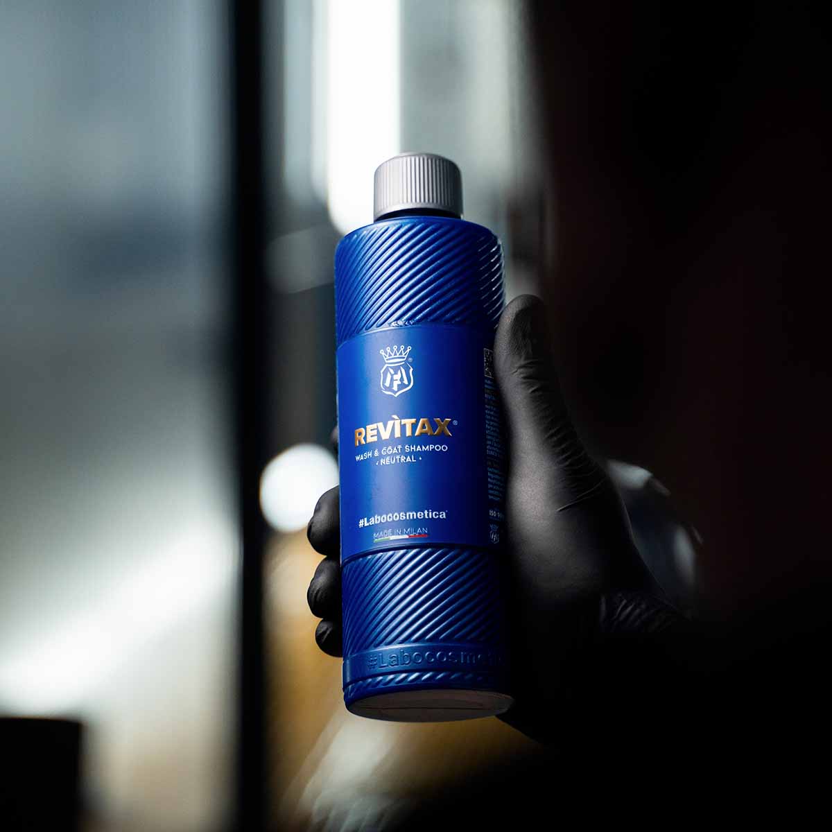 A hand in a black nitrile glove holding a blue bottle of Labocosmetica Revitax shampoo in a dark detailing studio.