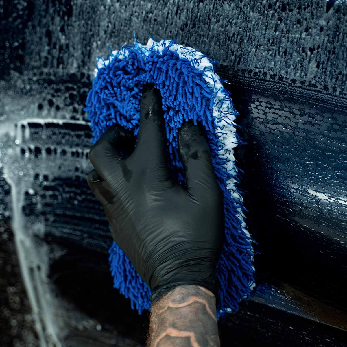 A gloved hand uses a sudsy blue microfiber wash mitt to wash a dark blue car panel with Labocosmetica Revitax shampoo.