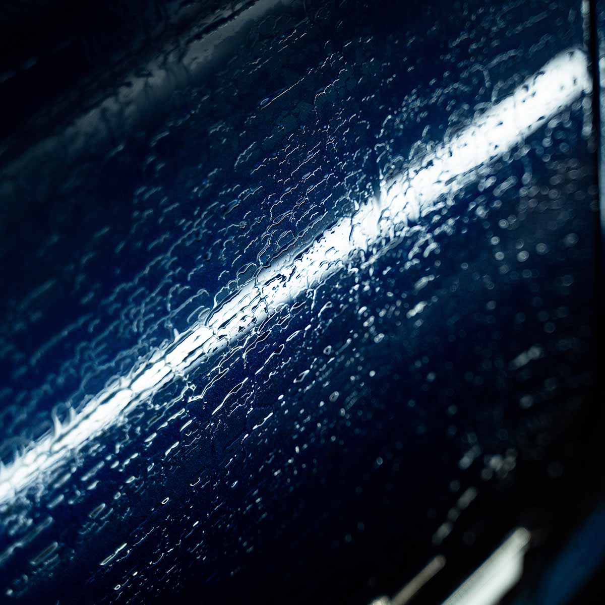 Close-up of water sheeting off a dark blue car panel with a bright light reflection, showing the protective properties of Labocosmetica Revitax.