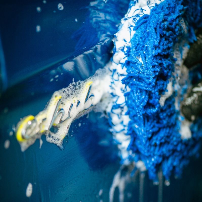 Close-up of a blue wash mitt cleaning the foamy blue trunk of a car, demonstrating safe washing with car shampoo.