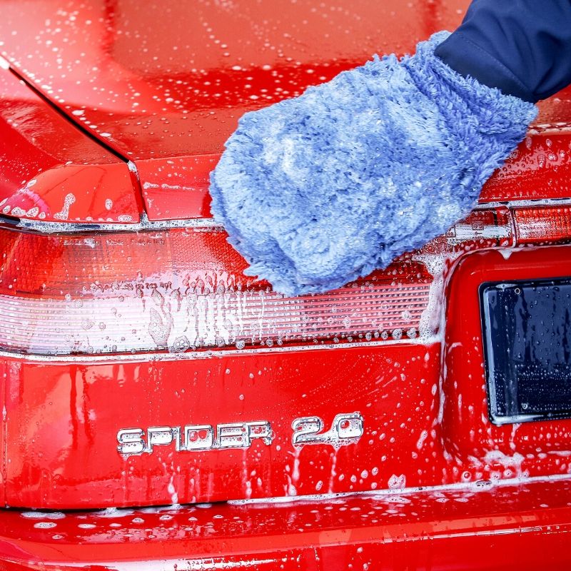 A hand wearing a blue wash mitt cleaning the foamy trunk of a red sports car, highlighting a safe, scratch-free car wash